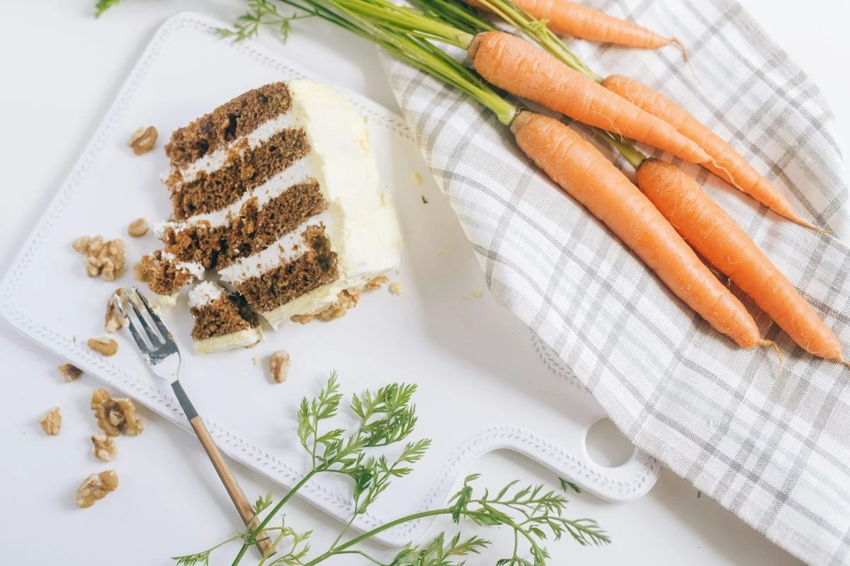 Ein Stück Karottenkuchen mit Cremefüllung, daneben Karotten und ein Geschirrtuch