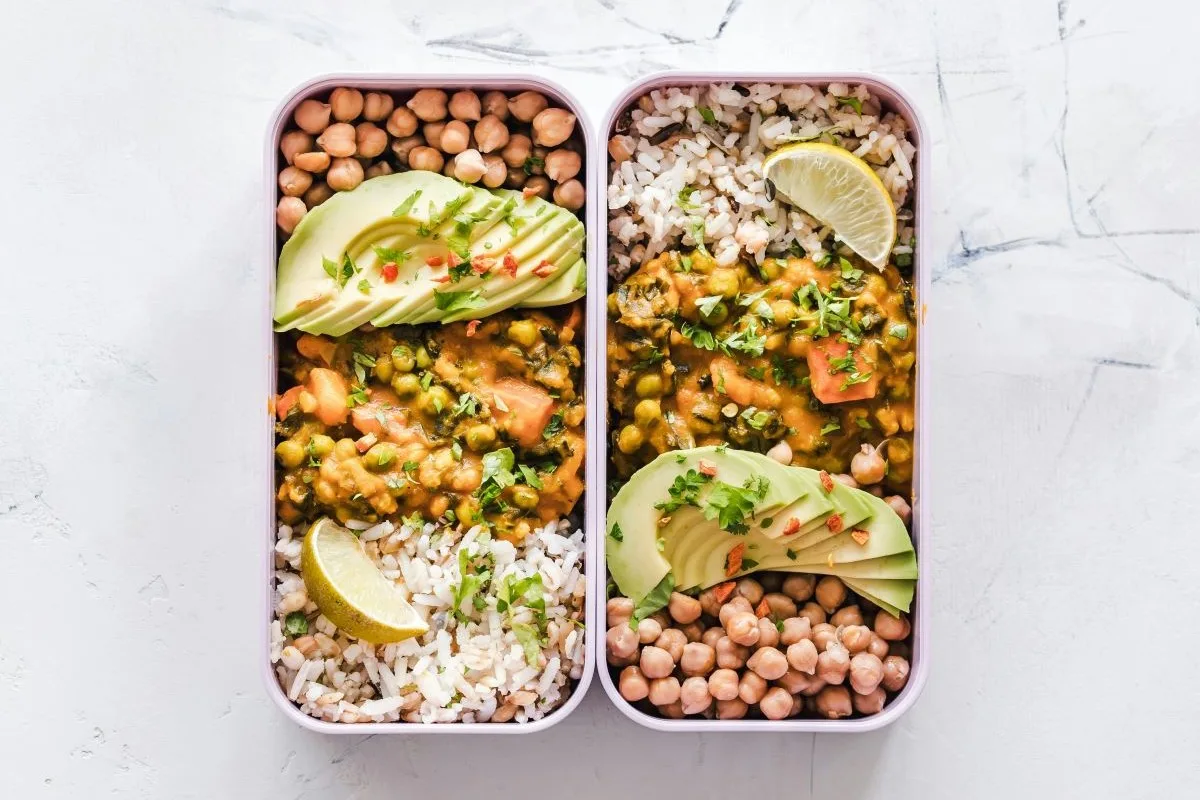 Zwei Jausenboxen, die mit Reis, Curry, Kichererbsen und Avocado gefüllt und mit einer Zitronenscheibe und Koriander garniert sind