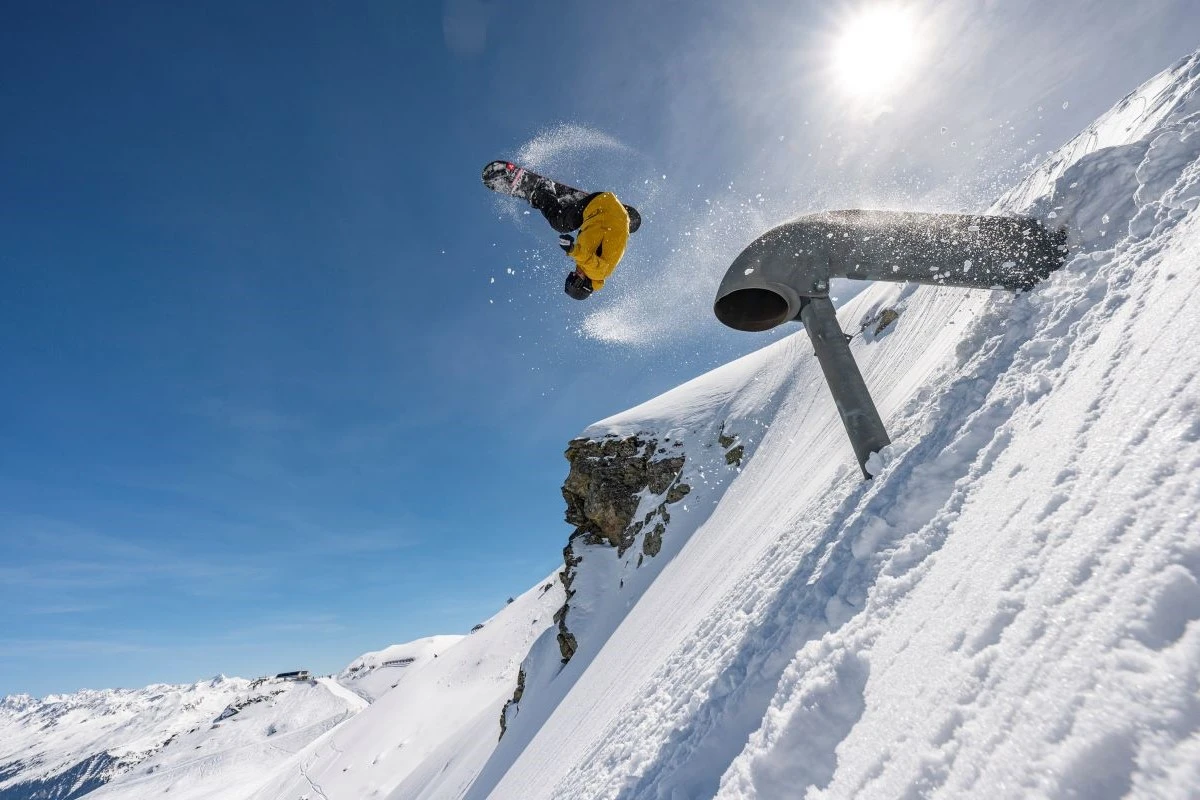 Thomas Feurstein fliegt mit seinem Snowboard kopfüber durch die Luft, der Hang ist schneebedeckt und im Hintergrund sind Berge zu sehen
